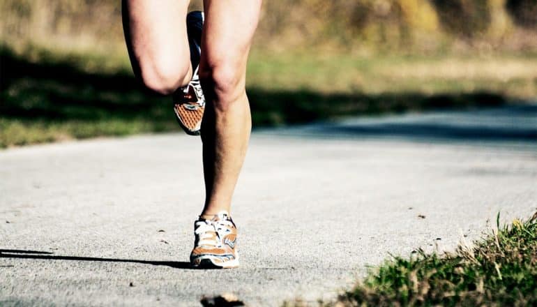 A person runs down a park path with grass on either side wearing orange running shoes, only their legs visible in the frame.