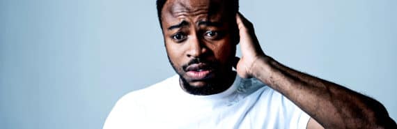 A man in a white t-shirt looks anxious or worried as he puts his hand to the back of his head