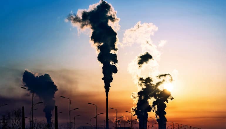 Dark clouds billow from a series of industrial smokestacks in silhouette against a setting sun and blue sky