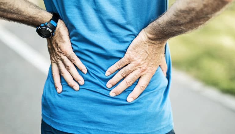 A man in a blue t-shirt holds his lower back in pain, with a path and grass in the background