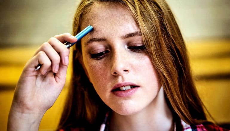 A college student holds a pen in her hand up to her forehead with a tired look on her face