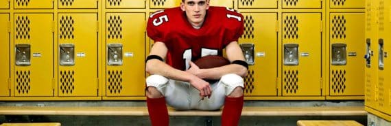 A young football player sits in a locker room in uniform with his helmet off