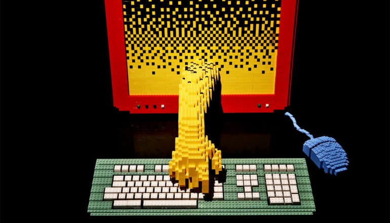 A Lego sculpture of a yellow hand reaching out of a computer screen and reaching for the keyboard, all against a black background