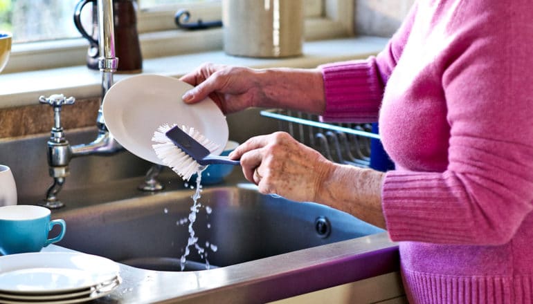 An older woman in a pink sweater washes a white dish with a scrubbing brush