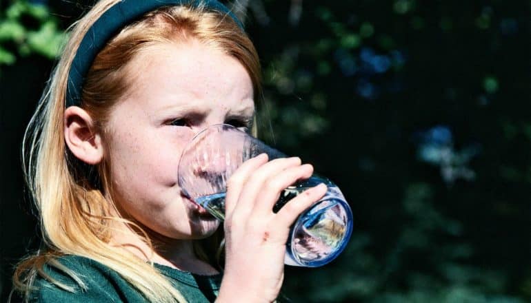 A little girl in green drinks a glass of water outside, with shadows and trees in the background