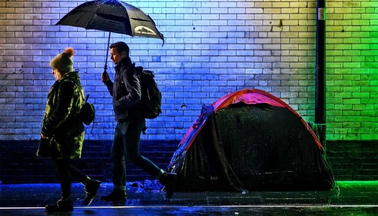 Two people walk past a tent on the sidewalk under a bridge, with blue and green lights illuminating the wet street