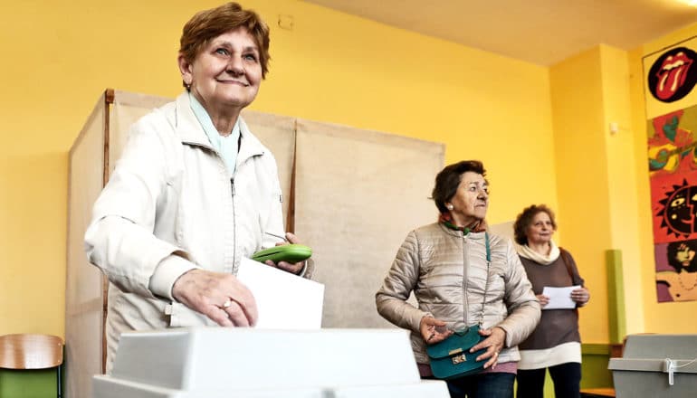 A woman smiles as she places her ballot in a box, with the voting booth and two other voters behind her in a yellow room