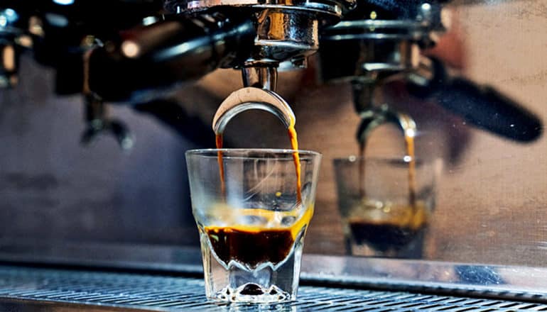 An espresso machine pours coffee into a small glass cup, which is reflected in the machine's metal