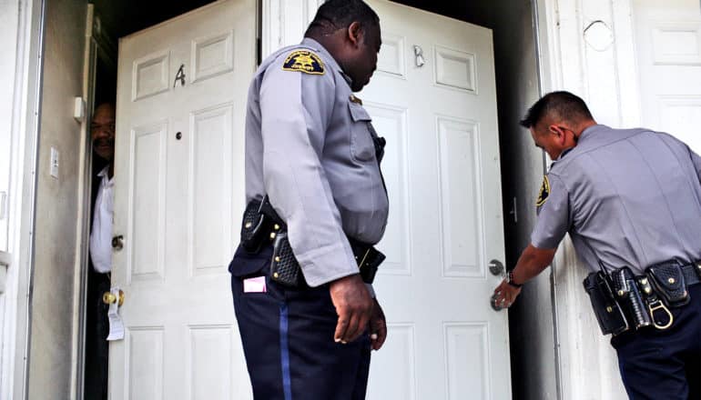 As police open the white door to an apartment, a neighbor opens his door and pokes his head out to see what's happening