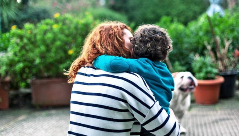 A mother in a white and black striped sweater hugs her son, who's wearing a teal hoodie, with plants and a bulldog in the background