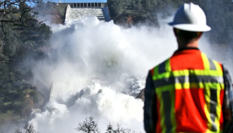 A worker in an orange safety vest and white hard hat with his back to the camera looks on as a cascade of white water rushes down the spillway built into a hill