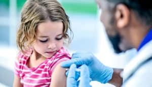 A little girl in a pink t-shirt gets a vaccine from a doctor wearing a white coat and blue gloves
