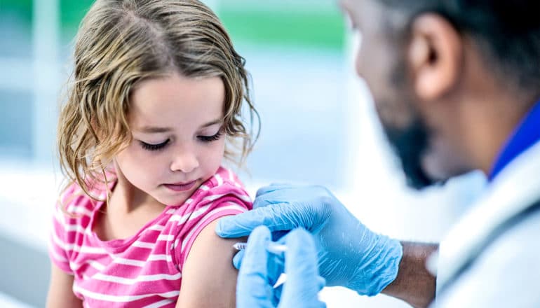 A little girl in a pink t-shirt gets a vaccine from a doctor wearing a white coat and blue gloves