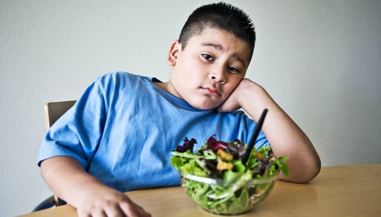A young boy in a blue shirt sits in front of a bowl of salad with a skeptical or disappointed look