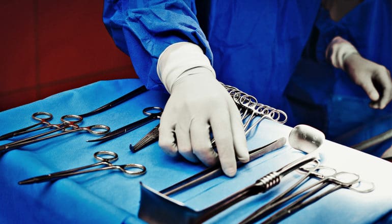 A surgeon with blue scrubs and white gloves reaches for a surgical tool sitting on a bright blue cloth