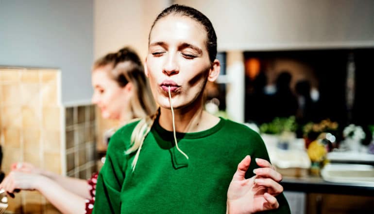 A woman in a green sweater sucks up a noodle with a satisfied look on her face and a person cooking in the background