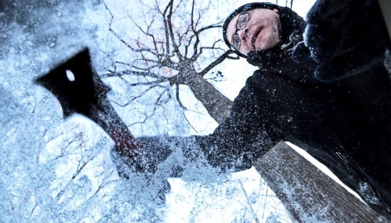 A man scrapes ice off his car windshield, viewed from inside the car looking up through the windshield