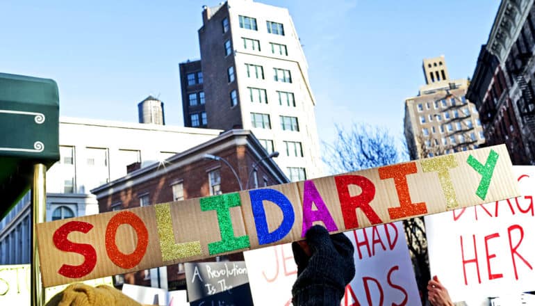 A protestor holds up a cardboard sign that reads "Solidarity" in rainbow-colored letters, against a backdrop of buildings and blue sky