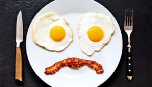 Two eggs and two pieces of bacon form a frowning face on a white plate, with a fork and knife on either side, against a dark table