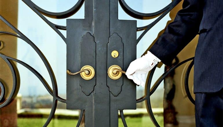 A white-gloved doorman reaches for the door knob on a glass and metal door