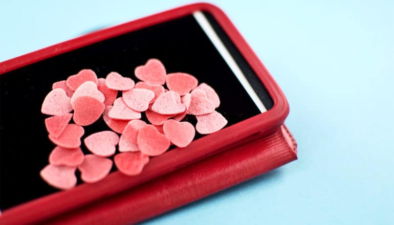 A phone in a red case sits on a blue background with many small hearts piling up on the black screen