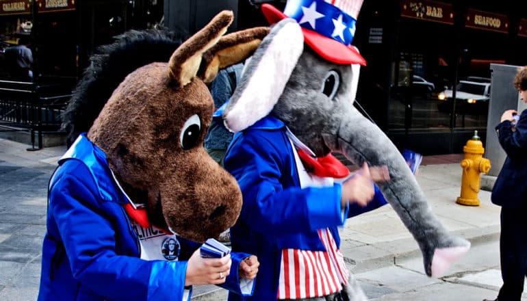 A person in a donkey costume and a person in an elephant costume walk down the street together, both with red, white, and blue suits