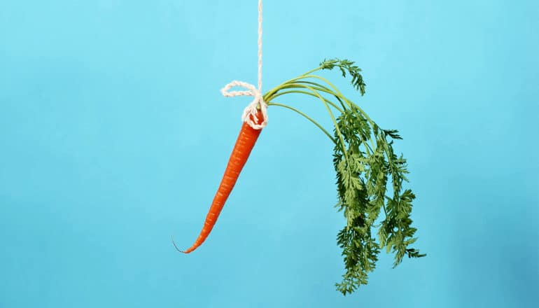 A bright orange carrot with green leaves hangs on a string against a blue background