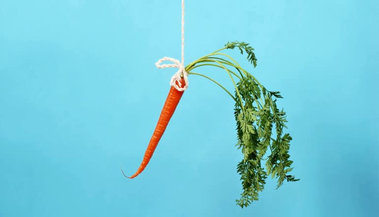 A bright orange carrot with green leaves hangs on a string against a blue background