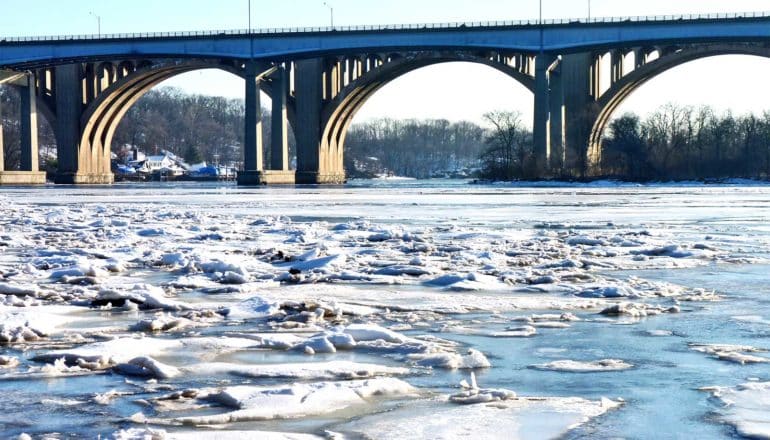 A partially-frozen river has chunks of ice floating in it. A bridge spans the river in the background, with a few houses along the distant shore