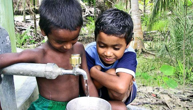 Two young boys look down at a faucet filling a water bucket with trees and plants in the background