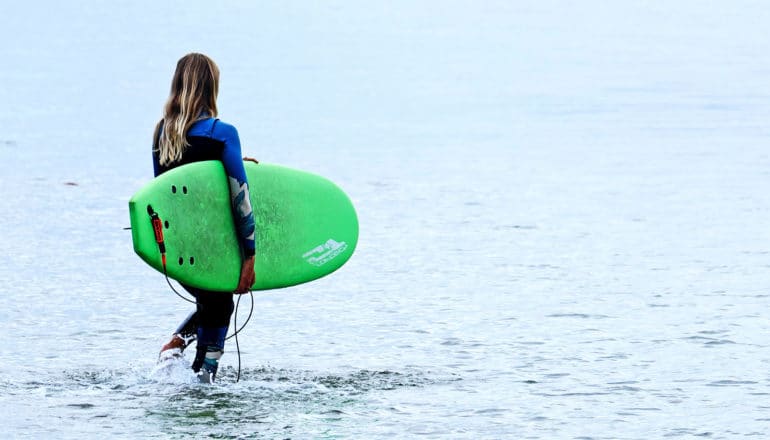 A woman in a wetsuit carries a green surfboard into the ocean
