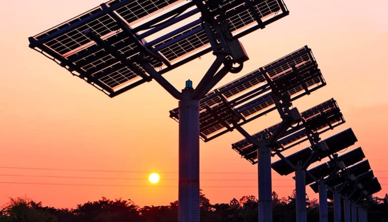 The sun sets in the distance, while a row of solar panels sit in the foreground