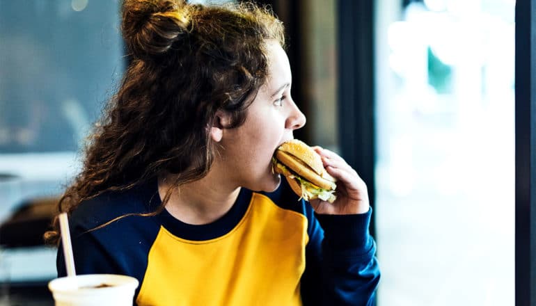 A teen girl in a blue and yellow eats a fast food burger while looking out the restaurant window
