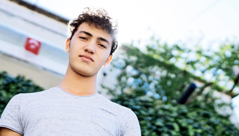 A teen boy looks down at the camera, with a roof-line, tree, and the sunny sky behind him