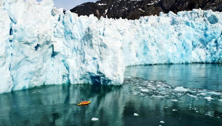 A yellow and orange robotic kayak floats near a massive wall of glacier ice