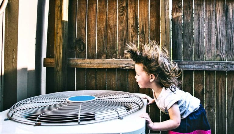 A young girl leans over an air conditioner unit as the air blows her hair up. Behind her and the AC unit, there's wooden fence