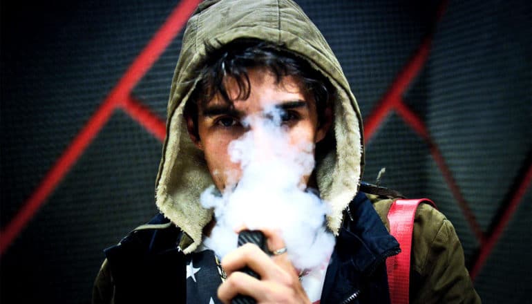 A teen in a hoodie is vaping, with a cloud rising in front of his face