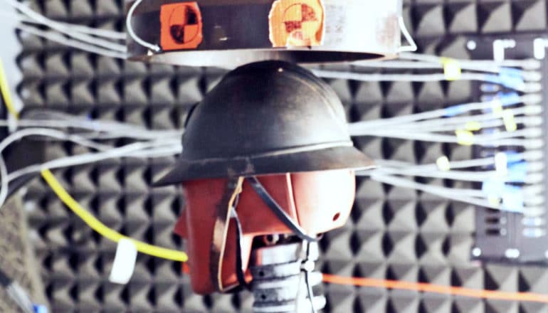 surrounded by insulating foam, dummy head wears helmet below metal tube with warning stickers