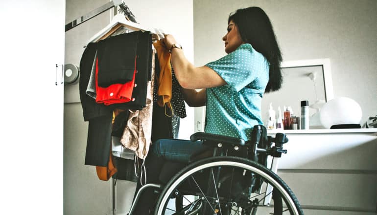 A woman in a blue shirt in a wheelchair is going through her clothes from her closet