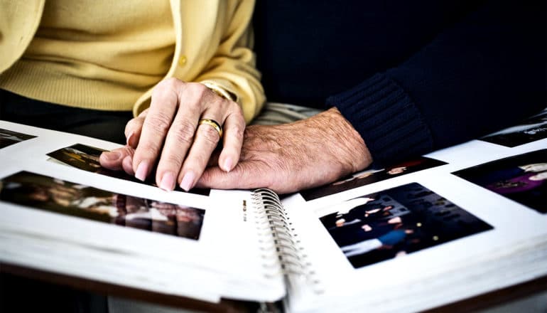 An older couple look at a photo album, with a woman in a yellow sweater putting her hand on his