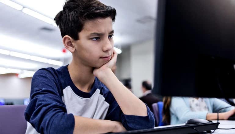 A student in a blue and white shirt looks at a computer screen in a computer lab in confusion, his head resting on his hand