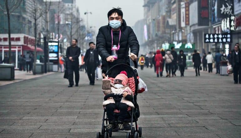 A man in a black jacket and blue medical mask pushes a stroller down a pedestrian walkway. In the stroller, a baby is wearing a medical mask as well