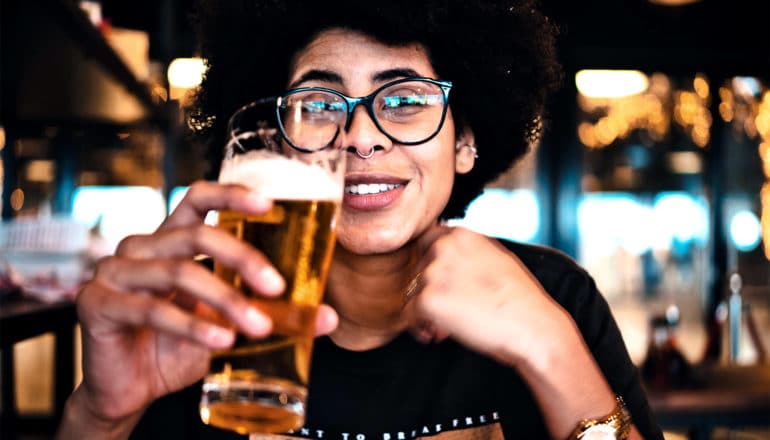 A young woman enjoys a beer in a bar, holding her glass while smiling across the table