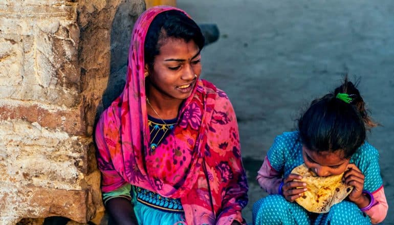 teen and child in colorful Indian clothes sit outdoors; child eats a chapati