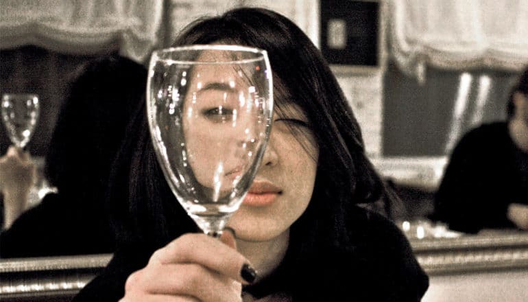 A young woman holds up an empty glass of wine to her eye as she sits in a restaurant