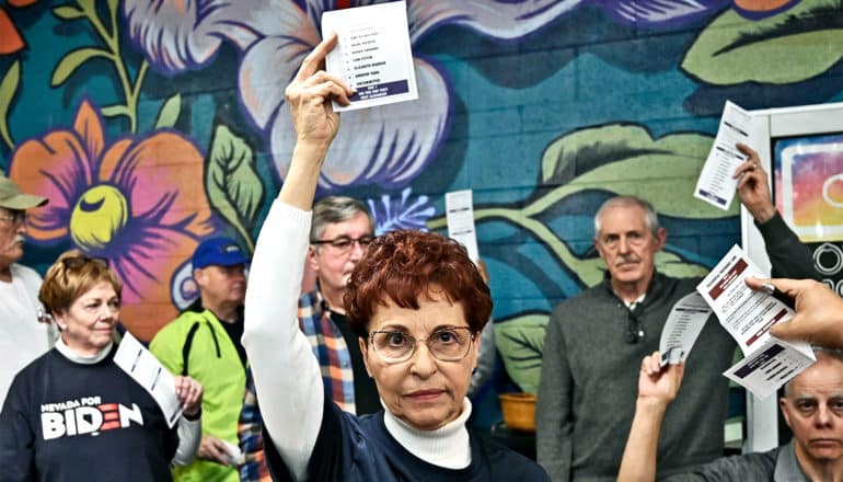 A woman with glasses and red hair holds up a piece of paper, with people behind her against a colorful painted wall