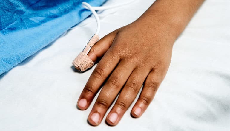 A child's hand lays on a hospital bed, with a piece of a blue hospital gown