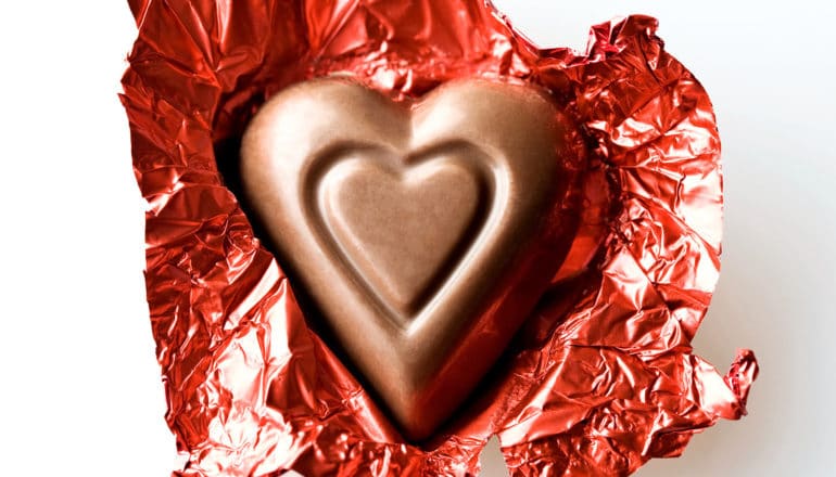 A heart-shaped piece of chocolate sits on its unwrapped red, shiny wrapper