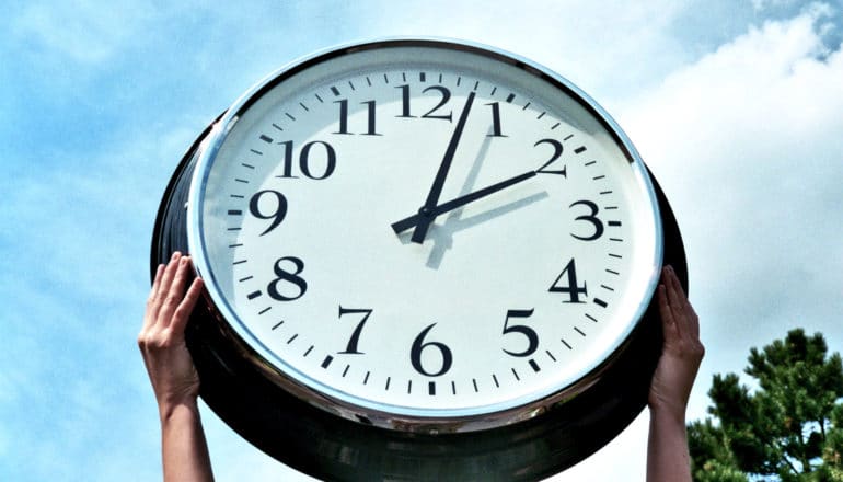 A pair of hands hold up a giant clock against a blue, cloudy sky