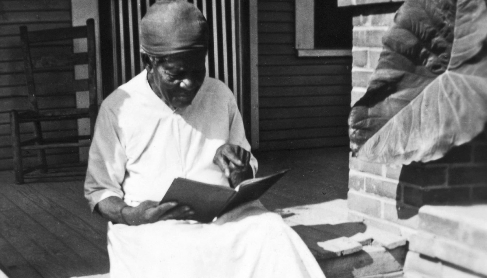 Mary Armstrong sits on a porch reading, while wearing a white dress and head wrap